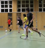 Several individuals are actively engaged in a game of indoor soccer within a gymnasium. The players are all in motion, with a focus on the ball located in the foreground. The setting is enclosed, featuring wooden panels and windows along the walls.