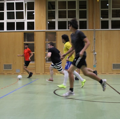 Several individuals are actively engaged in a game of indoor soccer within a gymnasium. The players are all in motion, with a focus on the ball located in the foreground. The setting is enclosed, featuring wooden panels and windows along the walls.