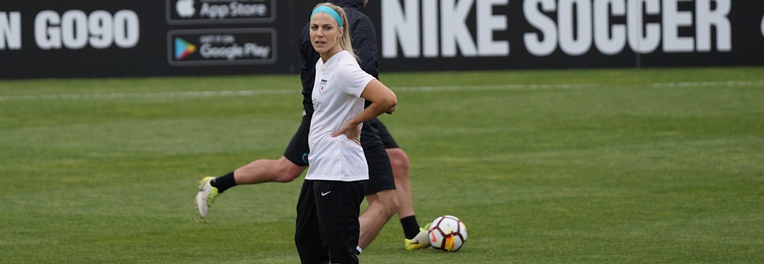 A person in sports attire stands on a soccer field, surrounded by several soccer balls and a carrier with bottles. The field is bordered by advertisements for Nike Soccer and app stores. Several people sit in bleachers in the background.