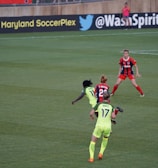 A soccer match taking place on a grassy field, featuring four players actively involved in the game. The players are wearing brightly colored jerseys, with one team in neon green and the other in red and blue. Advertising boards are visible in the background, indicating the location as Maryland SoccerPlex.