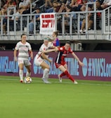 A competitive soccer match is underway with players vying for the ball near the sideline. The players wear different uniforms, indicating they are from opposing teams. The audience is visible in the stadium seating above, and there are visible advertisements and hashtags on the LED boards along the field.