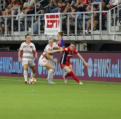 A competitive soccer match is underway with players vying for the ball near the sideline. The players wear different uniforms, indicating they are from opposing teams. The audience is visible in the stadium seating above, and there are visible advertisements and hashtags on the LED boards along the field.
