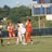 A group of female soccer players are actively engaged in a game on an outdoor field. The players are wearing red and white uniforms, with one goalkeeper in black. A soccer ball is airborne above them, indicating action and movement. A scoreboard in the background shows the score and other game details.