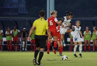Players on a soccer field are actively engaged in a game, with one player in red attempting to interact with the ball while another player in white jumps to intercept. A referee stands nearby in a yellow uniform. In the background, players in green bibs sit on the bench observing the action.