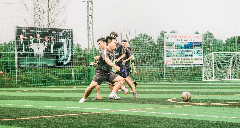 A group of young men are playing soccer on a sports field with artificial turf. The players are in motion, chasing after the ball, displaying a sense of dynamic action and teamwork. In the background, there is a fence with several large advertisement banners featuring logos and text.