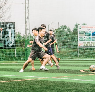 A group of young men are playing soccer on a sports field with artificial turf. The players are in motion, chasing after the ball, displaying a sense of dynamic action and teamwork. In the background, there is a fence with several large advertisement banners featuring logos and text.