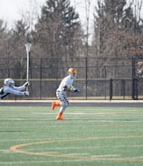 A group of athletes engaged in a sports game on a green field. One player is running forward with possession of the ball, wearing colorful socks and a checkered uniform, while another player in dark clothing dives to intercept or tackle. Other players are present in the background. Leafless trees and a metal fence surround the field.