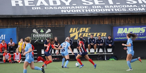 Players engage in a competitive soccer game on a field while others are seated on the bench watching attentively. The action is taking place in front of a wooden structure displaying multiple advertisements, including orthodontics, lacrosse, and field hockey.