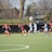 A group of female soccer players is actively engaged in a game on a green field, with two teams in different uniforms: light blue and dark blue with red accents. Nearby, a child wearing a yellow vest stands holding a soccer ball. The background features a black fence, leafless trees, and some park or neighborhood scenery.