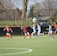 A group of female soccer players is actively engaged in a game on a green field, with two teams in different uniforms: light blue and dark blue with red accents. Nearby, a child wearing a yellow vest stands holding a soccer ball. The background features a black fence, leafless trees, and some park or neighborhood scenery.