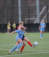 Players engaged in a competitive soccer match on a green field. One player in a blue uniform is actively kicking the ball while another player in a red and black uniform attempts to block. Two additional players in blue uniforms and another player in a red uniform are closely observing the action. The background shows others standing near the sideline.