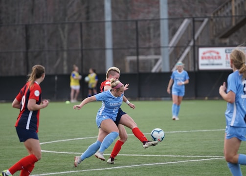 Players engaged in a competitive soccer match on a green field. One player in a blue uniform is actively kicking the ball while another player in a red and black uniform attempts to block. Two additional players in blue uniforms and another player in a red uniform are closely observing the action. The background shows others standing near the sideline.