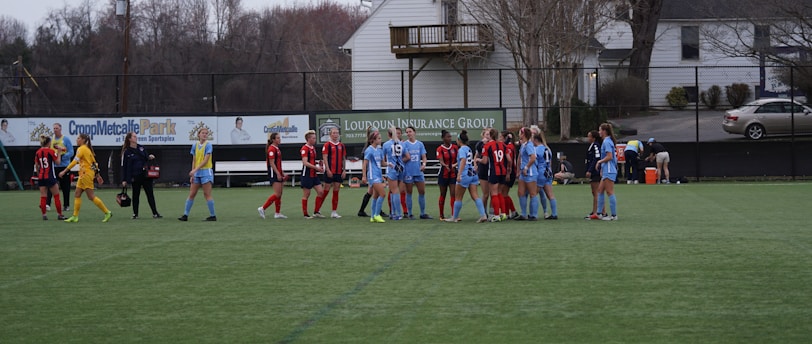 A group of female soccer players is standing on a green field. Two teams wearing different uniforms, one in red and black, and the other in blue, appear to be interacting before or after a game. There is a row of advertising boards and a residential area with trees in the background.