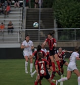 A group of female soccer players from two opposing teams are actively playing on a well-maintained green field. The players are wearing white and red kits, and they are jumping and reaching to head or kick a soccer ball in mid-air. The scene is dynamic and intense, capturing the action of a competitive match. Spectators can be seen seated in the background, watching the game with interest.