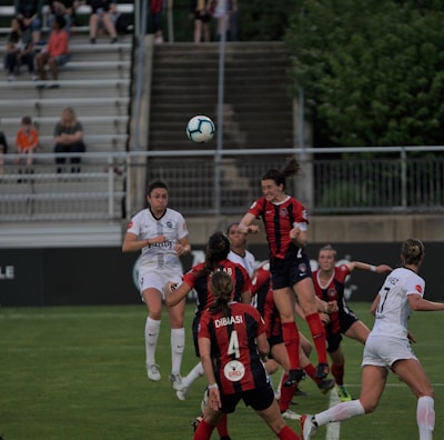 A group of female soccer players from two opposing teams are actively playing on a well-maintained green field. The players are wearing white and red kits, and they are jumping and reaching to head or kick a soccer ball in mid-air. The scene is dynamic and intense, capturing the action of a competitive match. Spectators can be seen seated in the background, watching the game with interest.