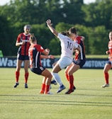 A group of women soccer players actively engaged in the game on a green soccer field. Three players in red and black uniforms are interacting with a player in a white uniform. The background features trees and some banners along the side of the field, indicating a professional soccer match environment.