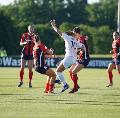 A group of women soccer players actively engaged in the game on a green soccer field. Three players in red and black uniforms are interacting with a player in a white uniform. The background features trees and some banners along the side of the field, indicating a professional soccer match environment.