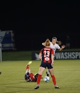 Several soccer players are engaged in an intense game on a field at night. One player in a red and blue uniform, marked with the number 23, is standing and observing the action. Another player is on the ground, wearing red shorts. A referee in a green jersey and black shorts is nearby. The background features a sign for 'WASHSPIRIT' and a partially visible goalpost.