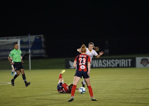 Several soccer players are engaged in an intense game on a field at night. One player in a red and blue uniform, marked with the number 23, is standing and observing the action. Another player is on the ground, wearing red shorts. A referee in a green jersey and black shorts is nearby. The background features a sign for 'WASHSPIRIT' and a partially visible goalpost.