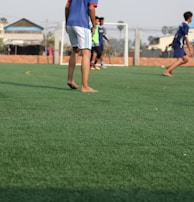 A group of young people are playing soccer on a green artificial turf field. The focus is on a few players in sportswear, with some of them appearing to be barefoot. A goalpost is visible in the background, along with a surrounding wire fence. Buildings and trees can be seen in the distance beyond the field.