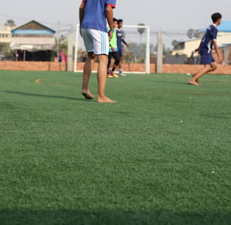 A group of young people are playing soccer on a green artificial turf field. The focus is on a few players in sportswear, with some of them appearing to be barefoot. A goalpost is visible in the background, along with a surrounding wire fence. Buildings and trees can be seen in the distance beyond the field.