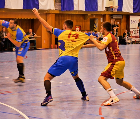 A dynamic indoor sports scene with players in motion on a court. Two athletes in blue and yellow uniforms are engaged in an intense handball game, with one player gripping the other's jersey. The background shows other players, several seated people, and advertisements on the walls.