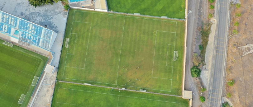 An aerial view of a sports complex featuring multiple green grass fields, including football pitches with marked goalposts. Adjacent to the fields is a blue-roofed building and a smaller area with a tennis or basketball court. Surrounding the complex, there's a road lined with trees and a dirt area with sparse vegetation.