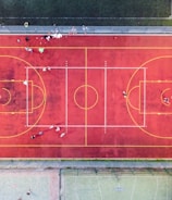 An aerial view of a sports court with a vibrant red surface and yellow lines marking different sections for basketball and soccer. Several people are present on the court, some engaged in activities, while others are standing along the perimeter. The court is enclosed by a metal fence and surrounded by grass on all sides.