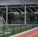 An outdoor sports facility featuring a running track and a soccer goal. The track is red, surrounded by a black metal fence, and the field is covered with artificial green turf. In the background, a basketball court is visible under a covered roof with metal supports. The sky is overcast, adding a moody atmosphere.