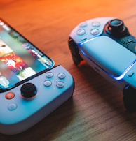A gaming scenario featuring a smartphone with a game interface next to two controllers on a wooden surface. The smartphone displays vibrant images and icons, while the controllers feature a sleek, modern design with buttons and joysticks prominently visible. Warm lighting enhances the scene.