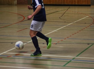 A person in a navy blue and white sports uniform is playing futsal in an indoor court. The individual is about to kick a colorful ball while wearing bright green and black shoes. The court is marked with various colored lines and has a wooden wall in the background.