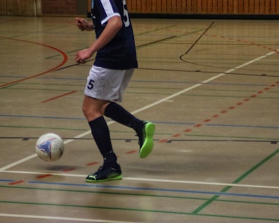 A person in a navy blue and white sports uniform is playing futsal in an indoor court. The individual is about to kick a colorful ball while wearing bright green and black shoes. The court is marked with various colored lines and has a wooden wall in the background.