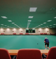 A large indoor sports facility featuring a green playing surface, similar to a bowling green. The ceiling is lined with numerous fluorescent lights, giving the space a bright yet serene atmosphere. Red chairs are arranged along the walls, and a scoreboard can be seen in the distance. A person is actively engaged in the game, bending forward as they release a ball, while inset a few spectators or participants are visible on the left side of the space.