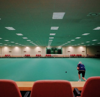 A large indoor sports facility featuring a green playing surface, similar to a bowling green. The ceiling is lined with numerous fluorescent lights, giving the space a bright yet serene atmosphere. Red chairs are arranged along the walls, and a scoreboard can be seen in the distance. A person is actively engaged in the game, bending forward as they release a ball, while inset a few spectators or participants are visible on the left side of the space.